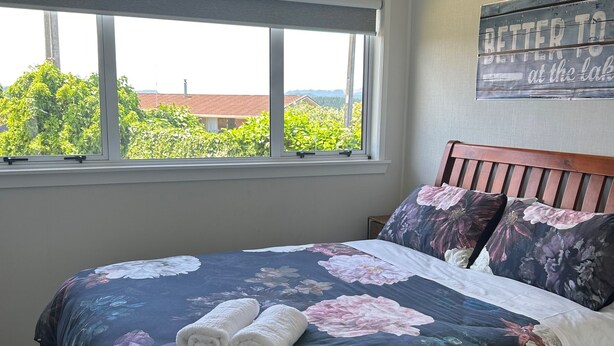 Ein Schlafzimmer mit großem Fenster, einem Bett mit Blumenmuster-Bettwäsche und Blick auf Grünflächen und Gebäude.