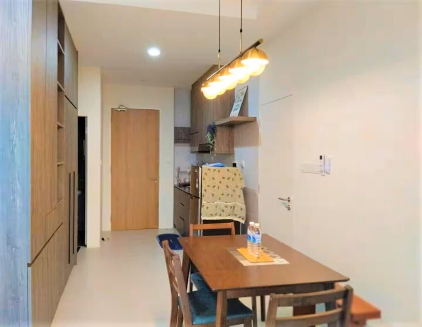 A dining area with a wooden table and chairs, a hanging light fixture, and a kitchen in the background.