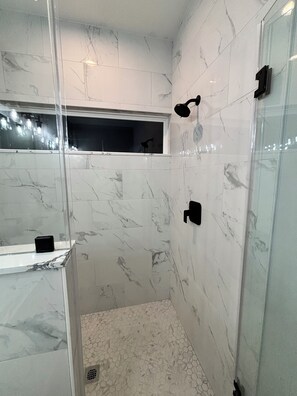 Ein modernes Badezimmer mit weißen Marmorfliesen, einer Duschkabine aus Glas und einem kleinen Fenster.