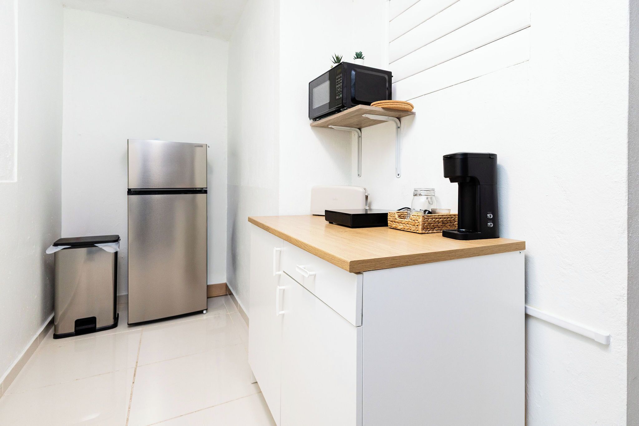 A compact kitchen with a stainless steel refrigerator, a microwave, a coffee maker, and a small trash bin.