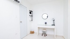 Une pièce blanche avec une porte, un bureau blanc avec un miroir, une petite plante en pot et un sac posé sur le sol.