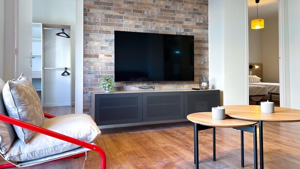 Un salon moderne avec un mur en briques, un téléviseur à écran plat, un fauteuil rouge, une table ronde en bois et une petite plante.