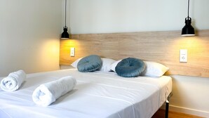 Un lit bien fait, avec du linge de lit blanc et deux serviettes enroulées, une tête de lit en bois et deux lampes noires fixées au mur.