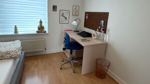 Une chambre à coucher comprenant un lit, un bureau, une chaise, un radiateur, une fenêtre avec des stores, des éléments de décoration murale et une petite plante.