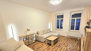A modern living room with a sectional sofa, a small wooden coffee table, and a side table with a vase. There are two windows with curtains, a radiator, and a wooden floor. - Modern 65m² apartment with parking space + balcony (Chemnitz)