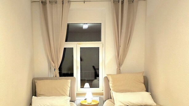 A room with two single beds, a small table with a lamp, a window with curtains, and a radiator. - Modern 65m² apartment with parking space + balcony (Chemnitz)