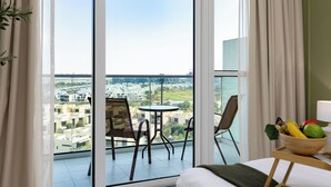 A hotel room with a balcony, a bed, a small table with a fruit bowl, and a view of the cityscape. - Maison Privee - Refined Studio w/ Top Facilities in Damac Hills (Dubai)