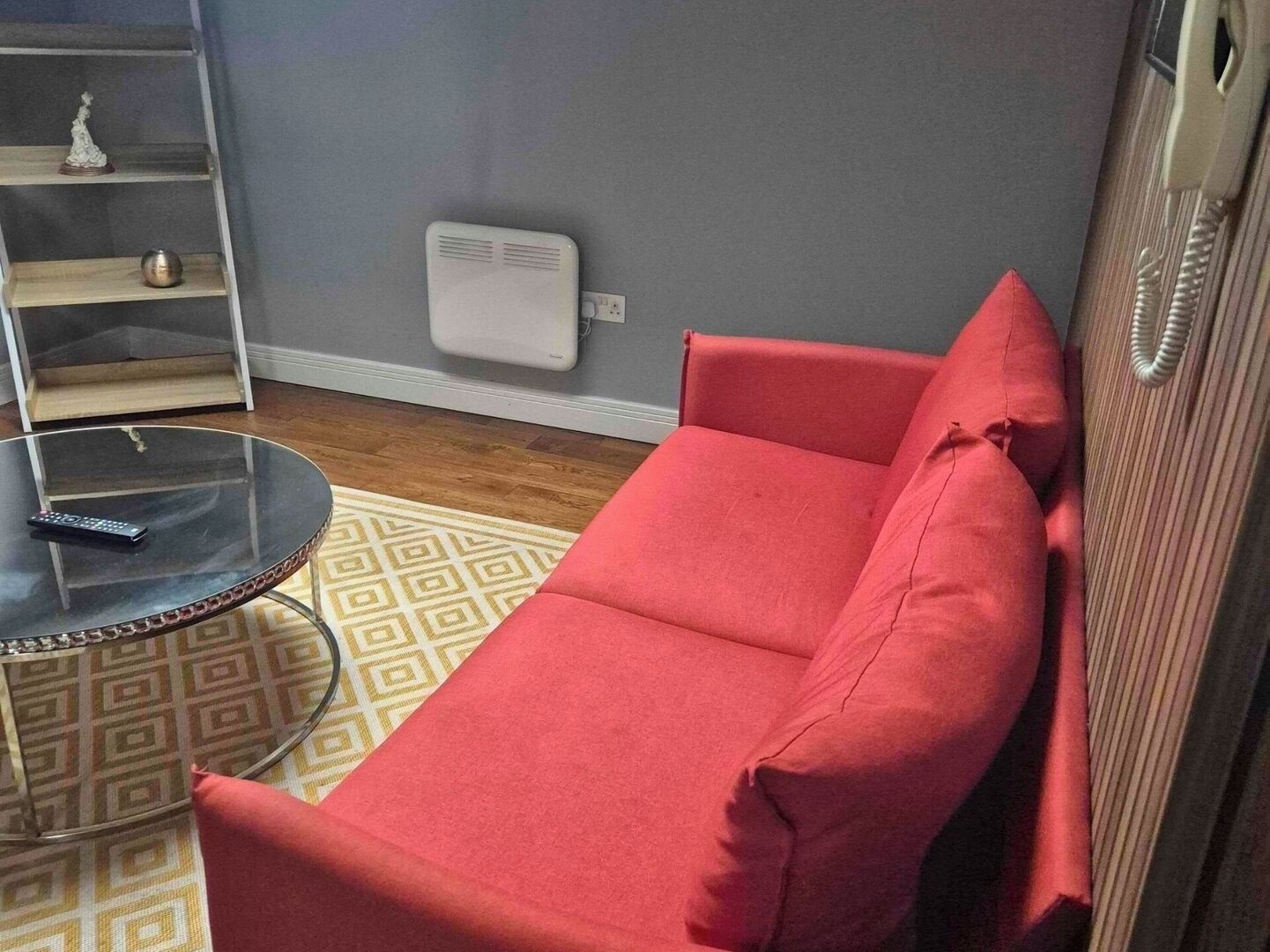 Un canapé rouge, une table basse en verre et un tapis à motifs dans une pièce dont le mur est gris et le plancher en bois.