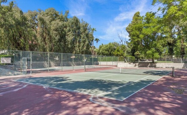 Un court de tennis en plein air, entouré d’une clôture métallique, d’arbres et bordé par un ciel dégagé.