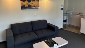 A modern room with a dark sofa, a small table with a remote control, and a painting of a landscape on the wall. - Charlies Motor Lodge (Balclutha)