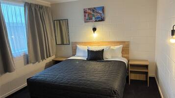 A hotel room with a large bed, a nightstand, a wall-mounted lamp, a window with curtains, and a painting on the wall.