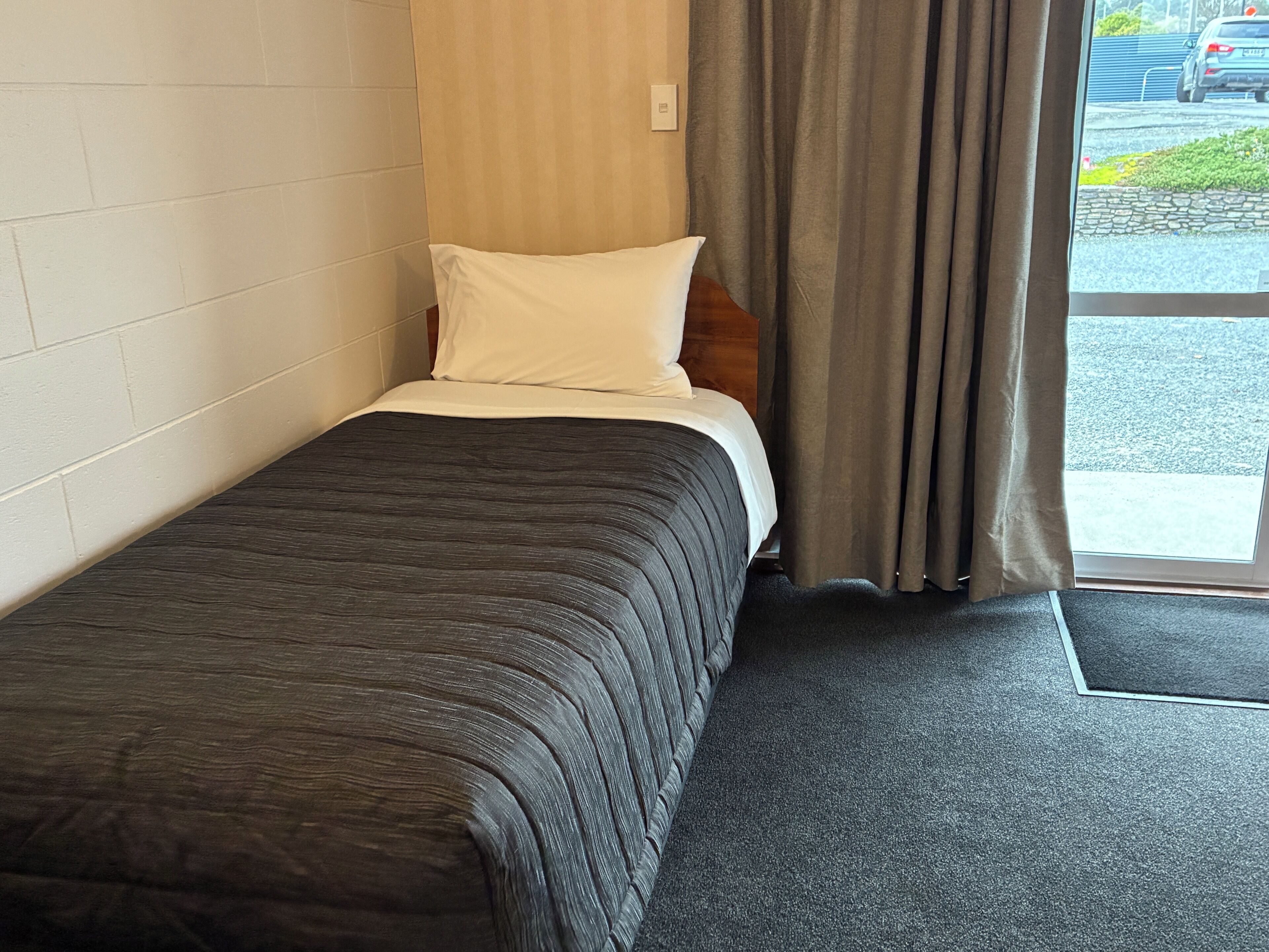 A single bed with a dark quilt, a white pillow, and a wooden headboard in a room with a window and curtains.