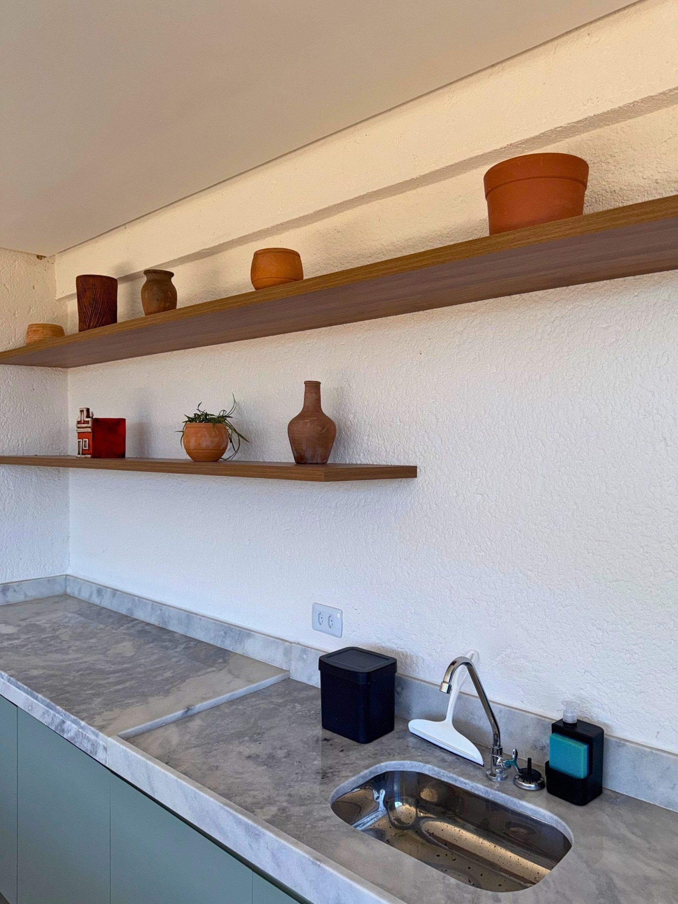 A kitchen with a marble countertop, a stainless steel sink, wooden shelves with pottery, and a small potted plant.