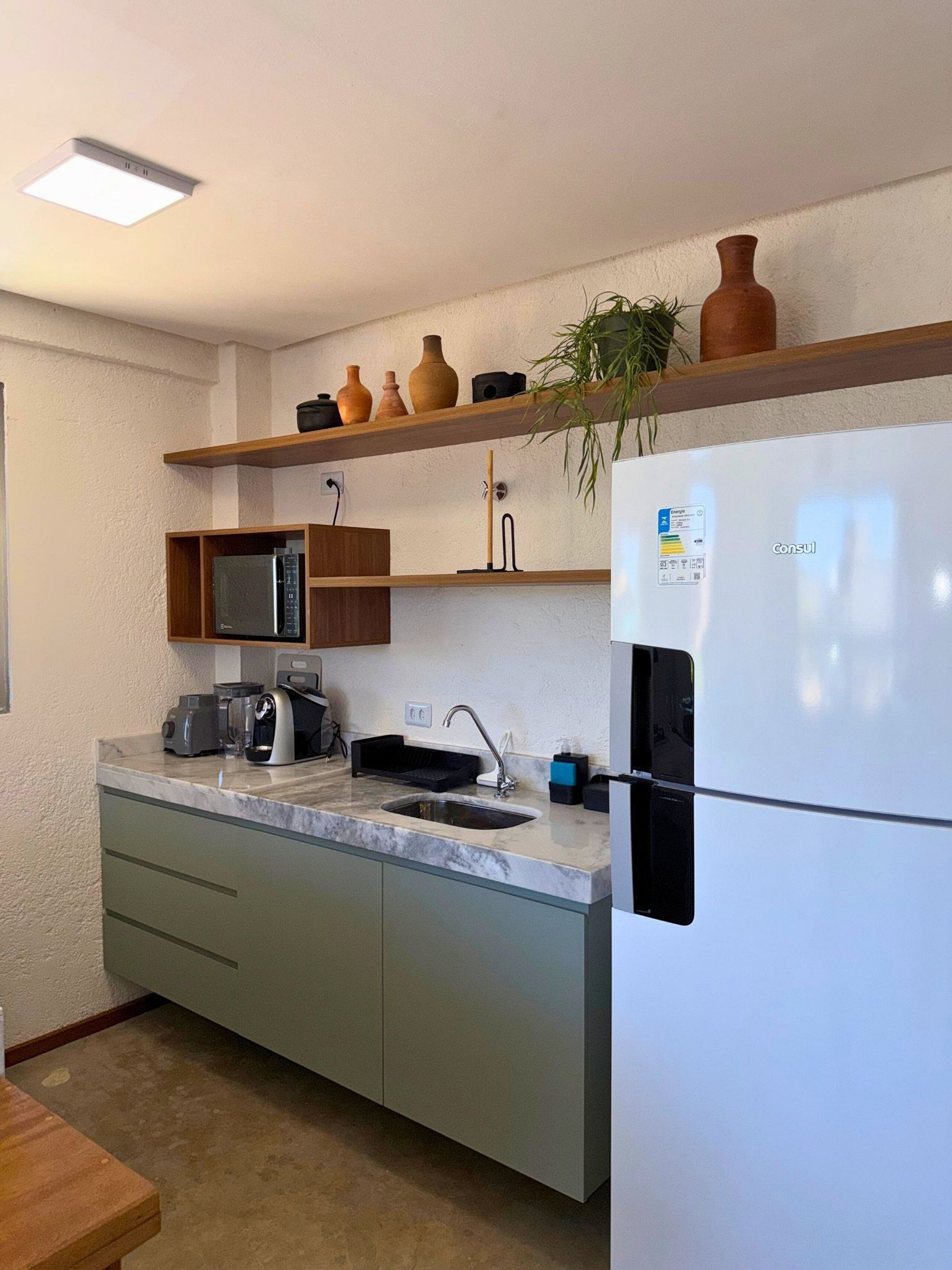 Una cocina con un refrigerador blanco, un microondas, un lavamanos y una cafetera. Hay repisas con adornos y una planta.