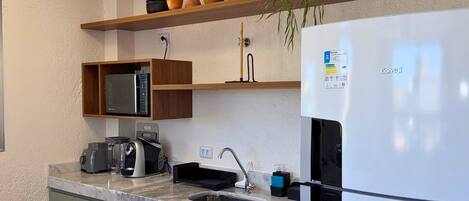 Una cocina con un refrigerador blanco, un microondas, un lavamanos y una cafetera. Hay repisas con adornos y una planta.