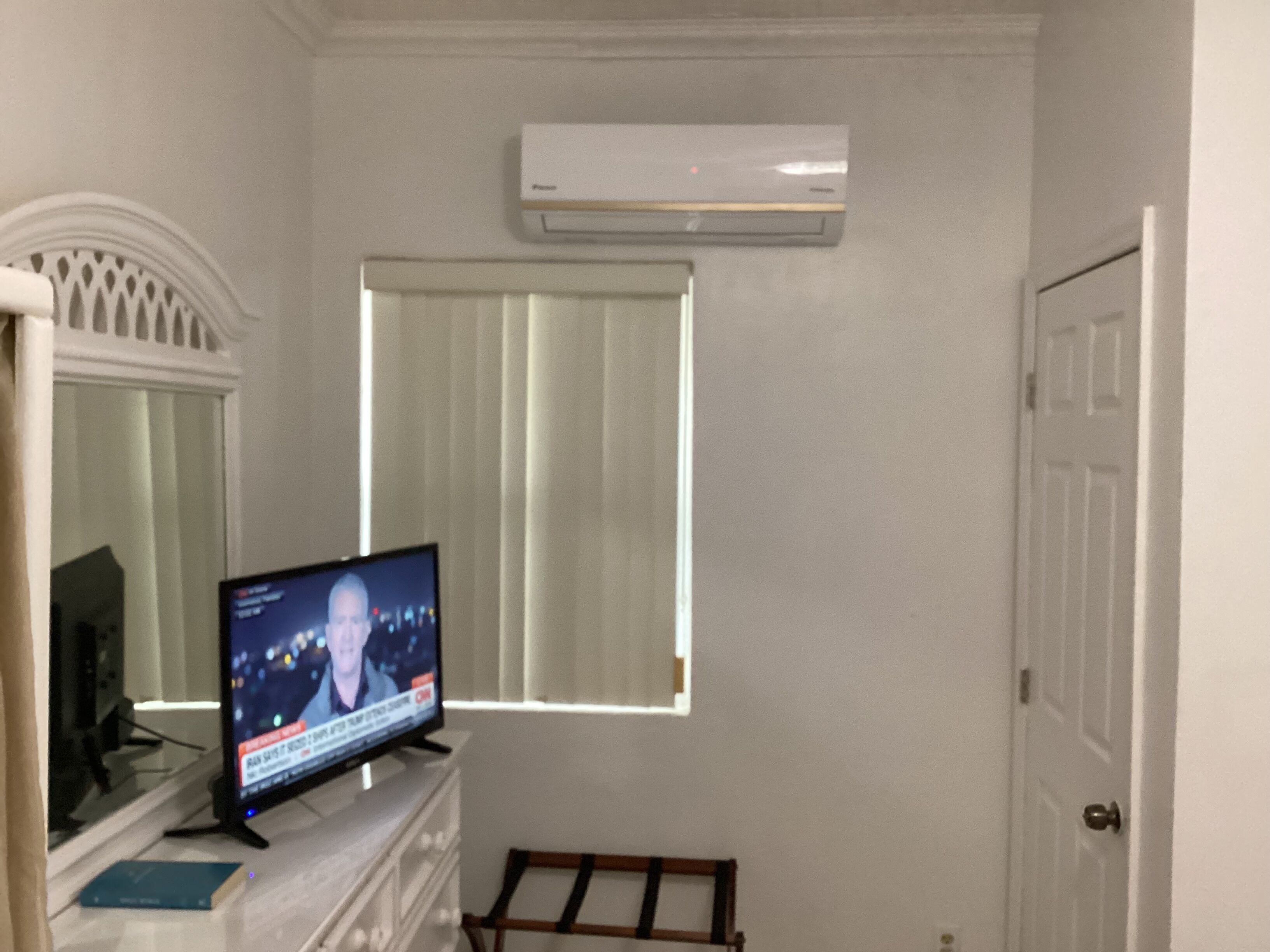 Una habitación con una televisión en una cómoda, una ventana con persianas, aire acondicionado y una puerta.