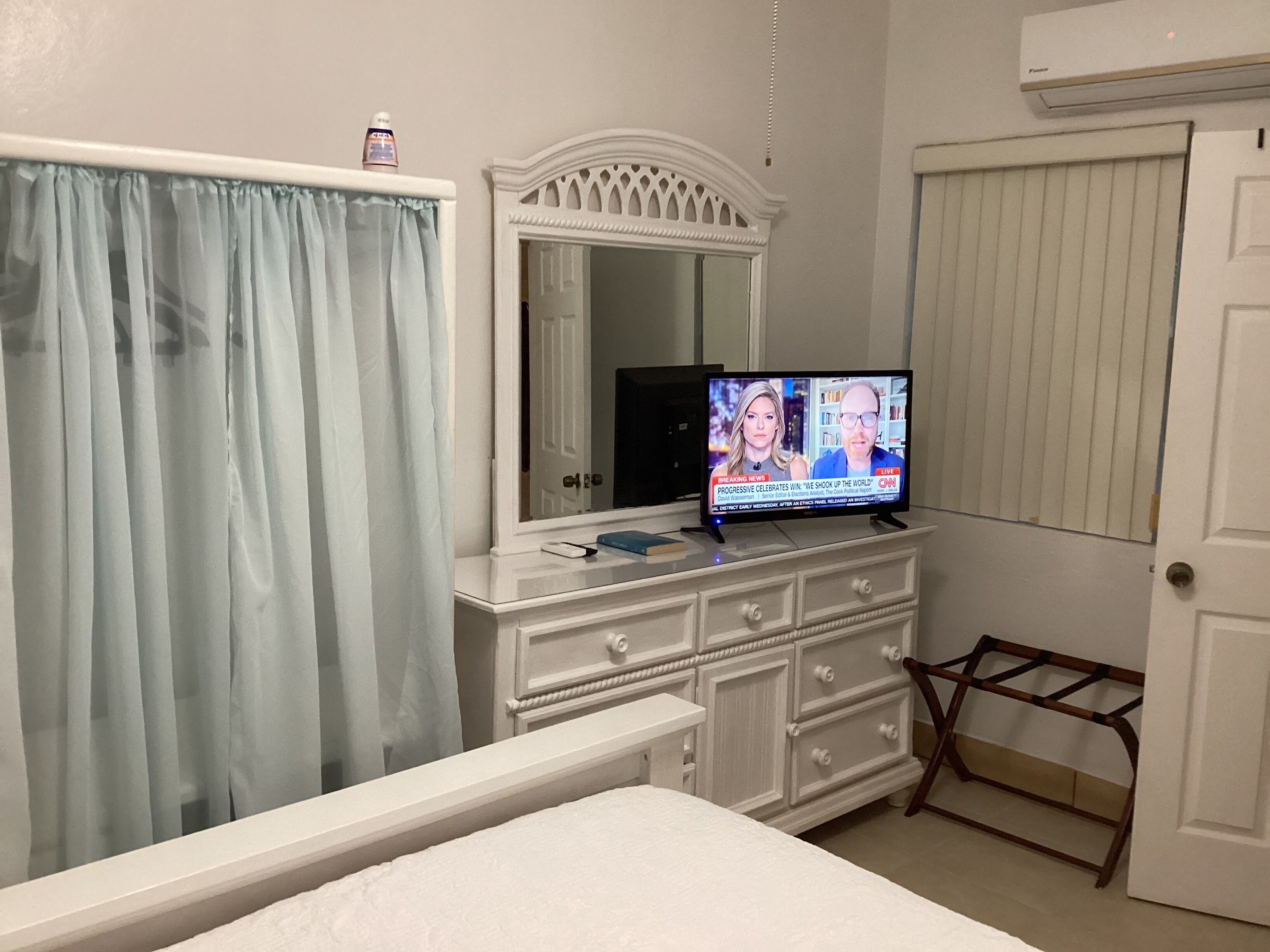A bedroom with a bed, a dresser, a television displaying a news program, a mirror, and a window with curtains.