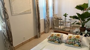 A neatly made bed with two decorative pillows, a small table with a plant, and a chair in a room with patterned curtains and a tiled floor.