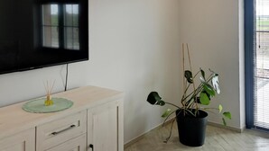 A room with a flat-screen TV mounted on the wall, a wooden cabinet with drawers, a potted plant, and a window with blinds.