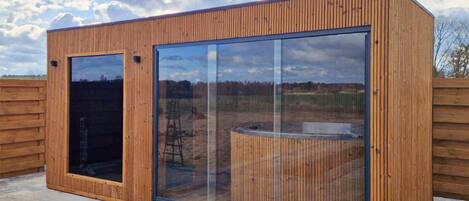 Une structure moderne en bois, dotée de grandes fenêtres en verre, qui ressemble probablement à un sauna ou à un espace de vie extérieur, et qui se détache sur un paysage rural.