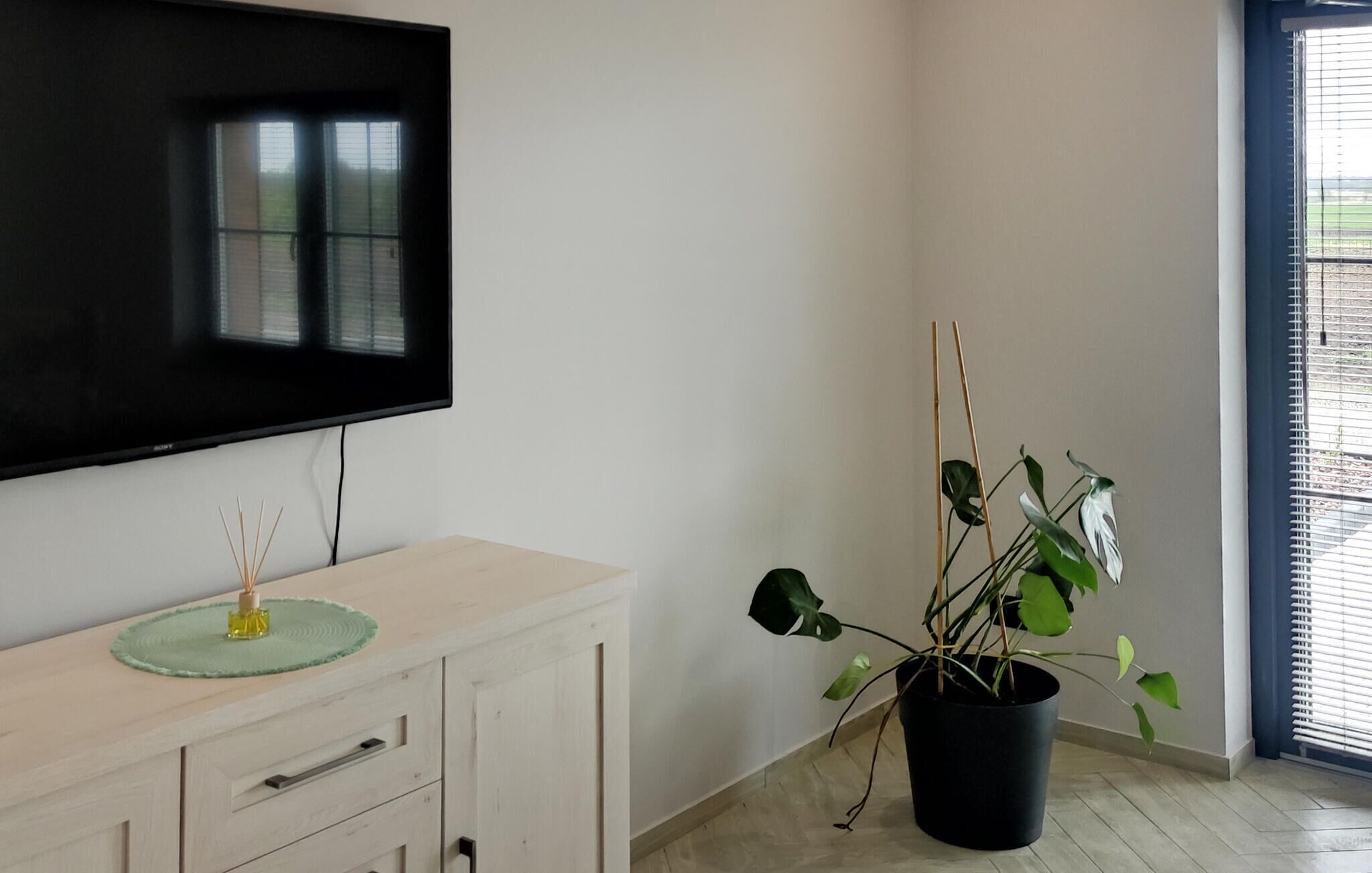 A room with a flat-screen TV mounted on the wall, a wooden cabinet with drawers, a potted plant, and a window with blinds.