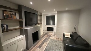 Un salon moderne avec un téléviseur à écran plat fixé au-dessus d’une cheminée, un canapé et une fenêtre équipée de stores.