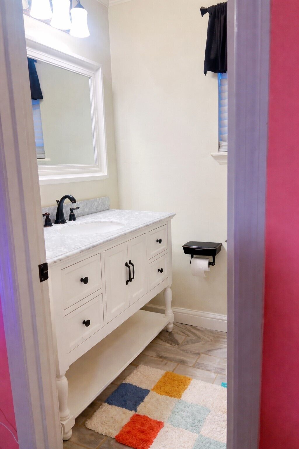 A bathroom with a white vanity, a mirror, a black towel rack, and a colorful rug.