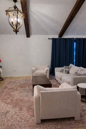 Ein geräumiges Wohnzimmer mit hoher Decke, einem Holzbalken, einem gemusterten Teppich und einem dunkelblauen Vorhang.