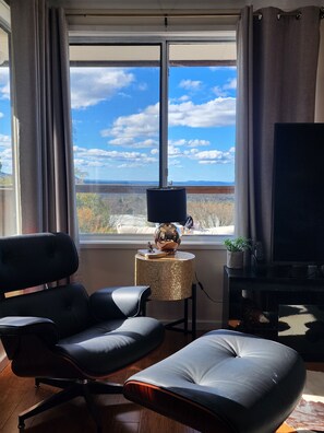 A modern living room with a large window, a black leather sofa, a small round side table with a lamp, and a flat-screen TV. - Luxury Panorama Spa & Sauna Retreat with Blue Mountains Views (Wentworth Falls)