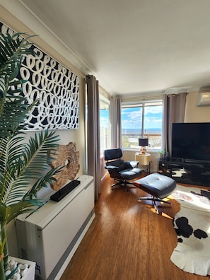 A modern living room with a large window, a flat-screen TV, a comfortable seating area, and a decorative wall piece. - Luxury Panorama Spa & Sauna Retreat with Blue Mountains Views (Wentworth Falls)