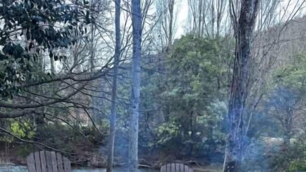 A circular fire pit with burning logs and two wooden chairs around it, set in a natural outdoor environment with trees and a river in the background. - Lazy Daze River Retreat (Kevington)