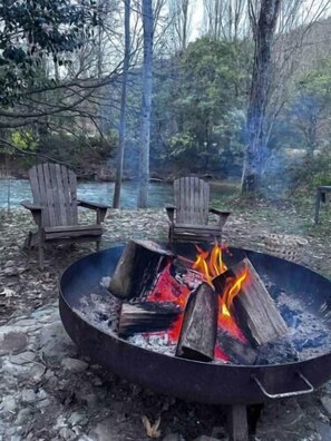 Un foyer circulaire, avec des bûches qui brûlent, entouré de deux chaises en bois, installé dans un cadre naturel en plein air, avec des arbres et une rivière en arrière-plan.