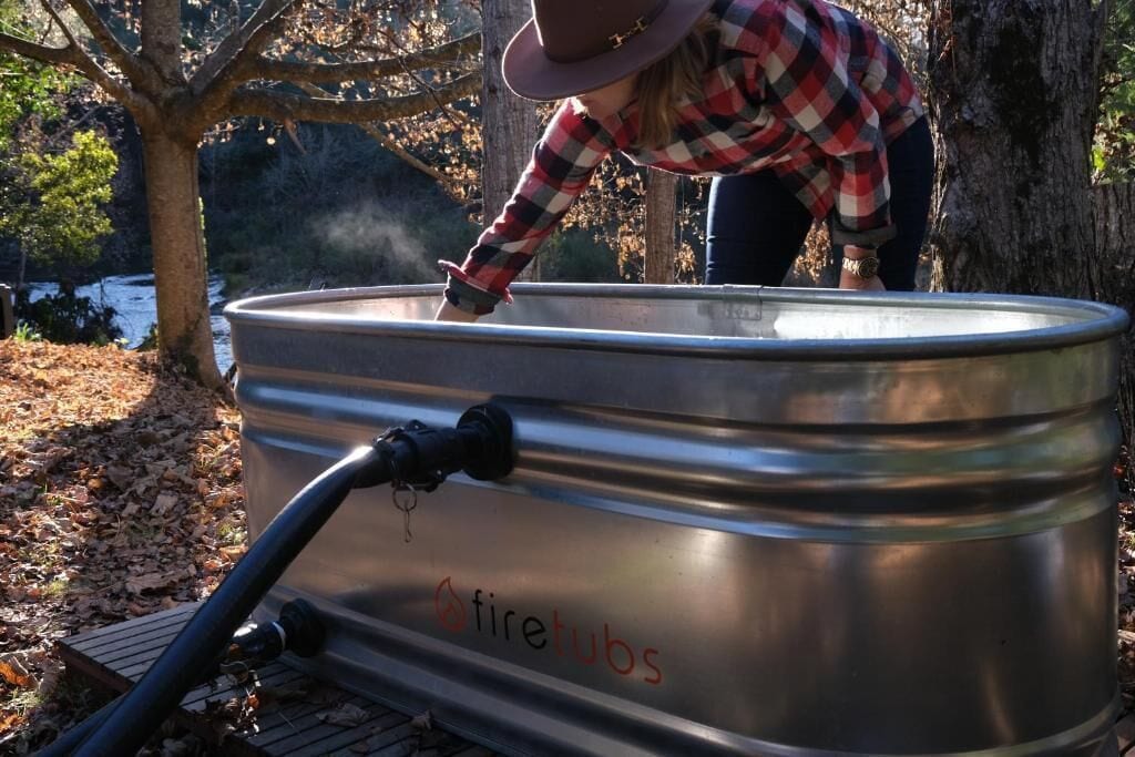 A stainless steel container with the word "firetubs" on it, outdoors with trees and a leaf-covered ground.