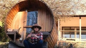 A person sitting outside a wooden barrel-shaped structure with a window and a door. - Lazy Daze River Retreat (Kevington)