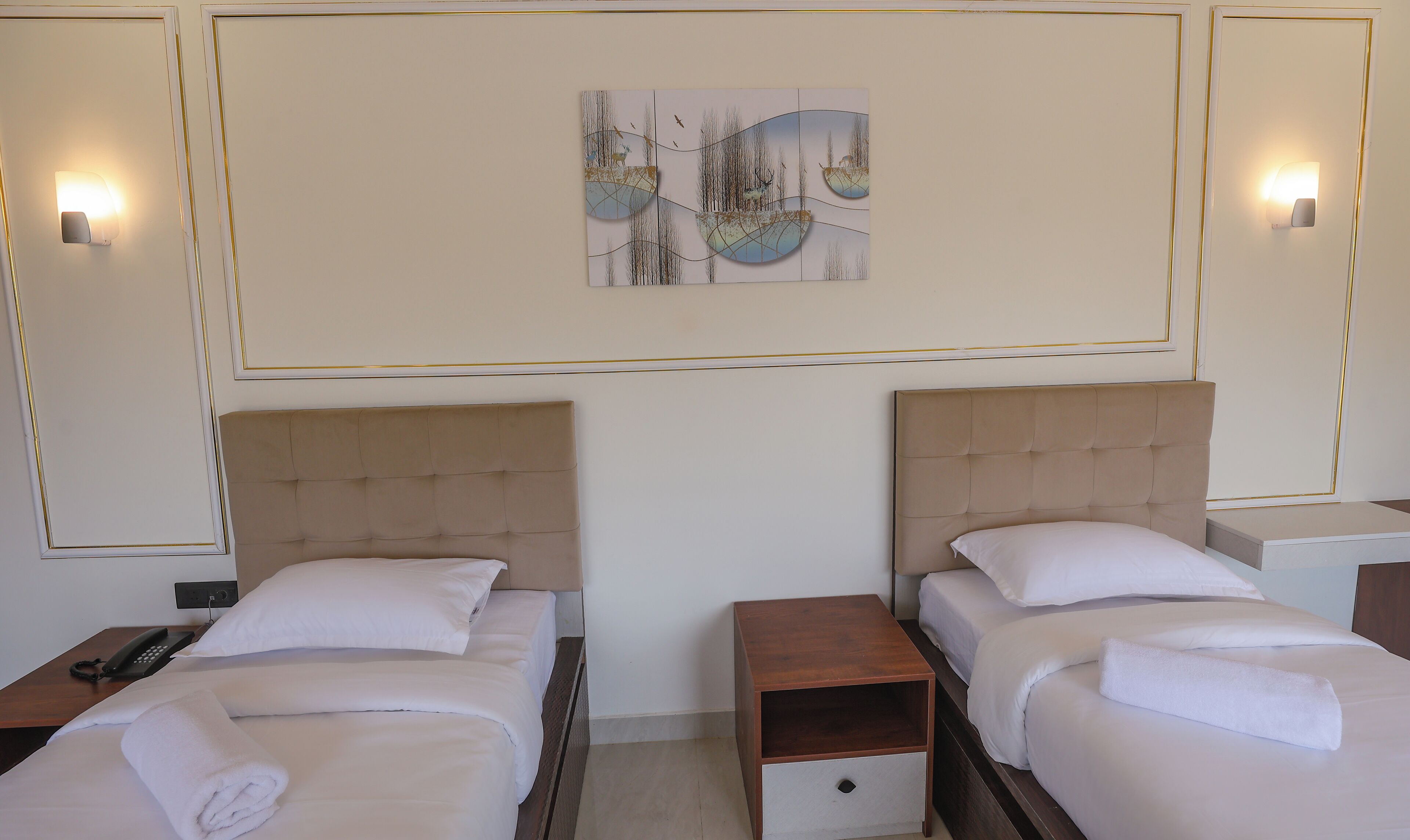 Quarto de hotel com duas camas, cada uma com cabeceira, mesa de cabeceira e luminária fixa na parede.
