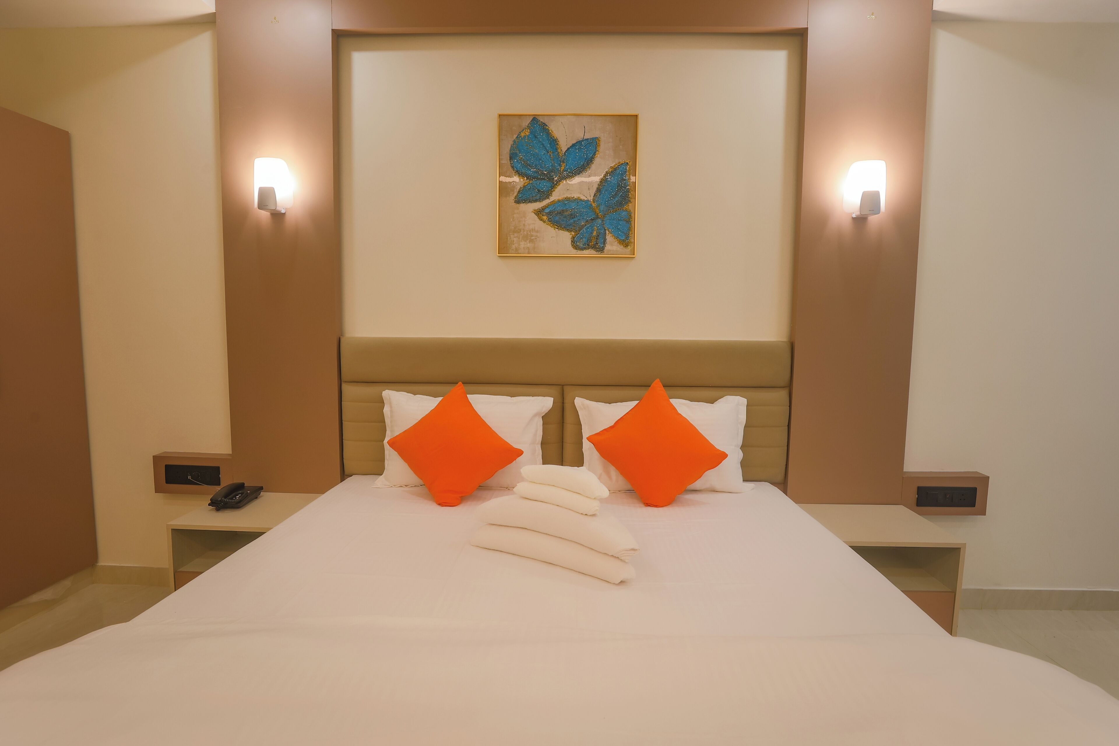 Ein Hotelzimmer mit einem großen Bett, zwei orangefarbenen Kissen, einer weißen Bettdecke, einem Wandbild von blauen Schmetterlingen und Nachttischlampen.