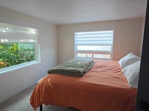 A bedroom with a bed, two windows with blinds, and a view of greenery outside.