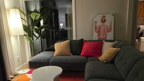 Una sala de estar moderna con un sofá seccional gris, una mesa de centro blanca y una alfombra roja. Se ve una planta grande y un cuadro en la pared.