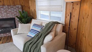 Una sala de estar acogedora con chimenea, un sofá con cojines, una lámpara de pie y una ventana con persianas.
