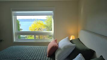 A bedroom with a bed, pillows, a nightstand, a lamp, and a window with a view of a body of water and trees.