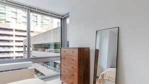 A bedroom with a large window, a wooden dresser, and a mirror. - Blueground | Long Island City, pool, nr Gantry Plaza (New York)