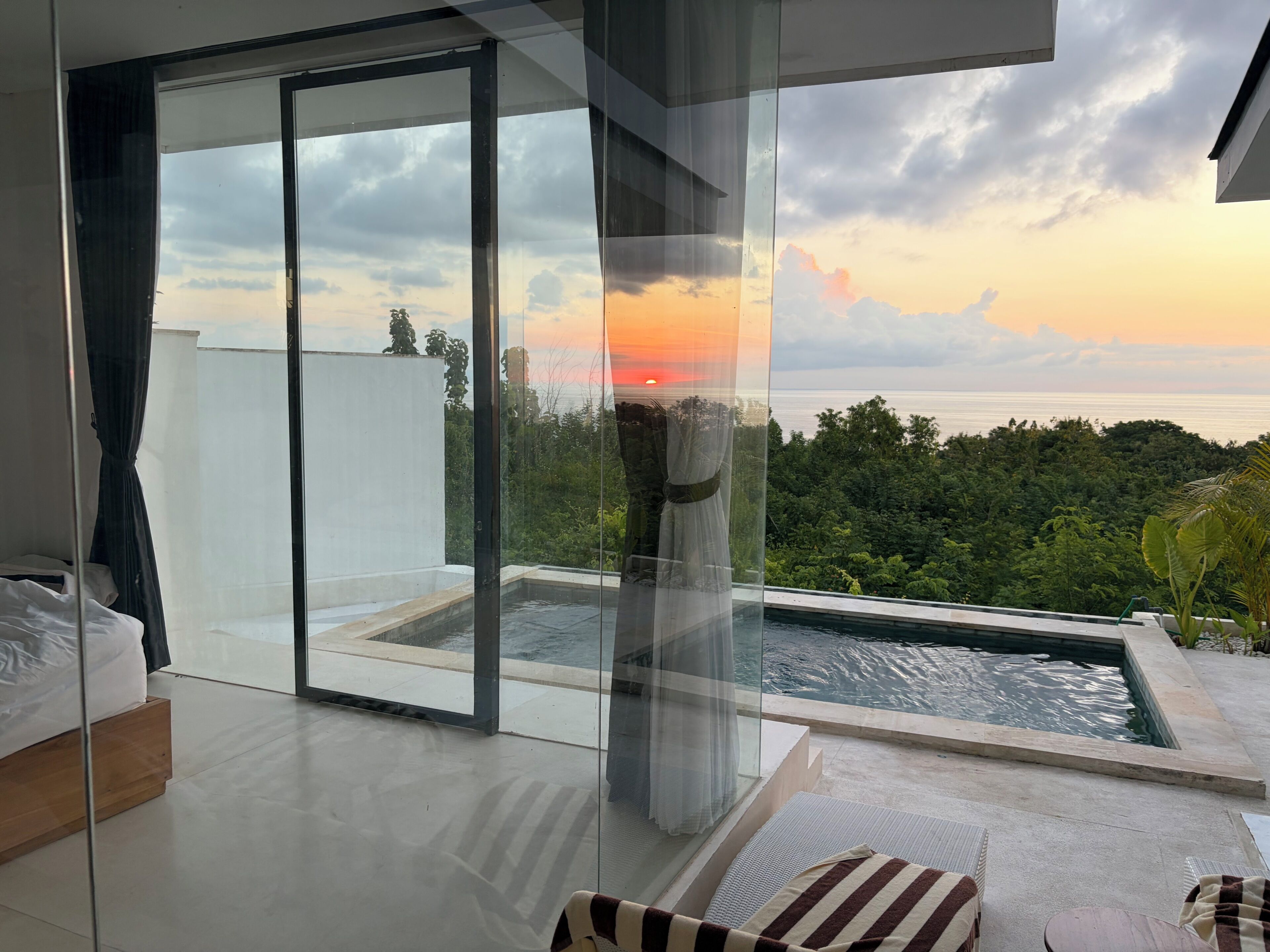 A modern room with a glass door leading to a balcony with a pool and a view of the ocean at sunset.