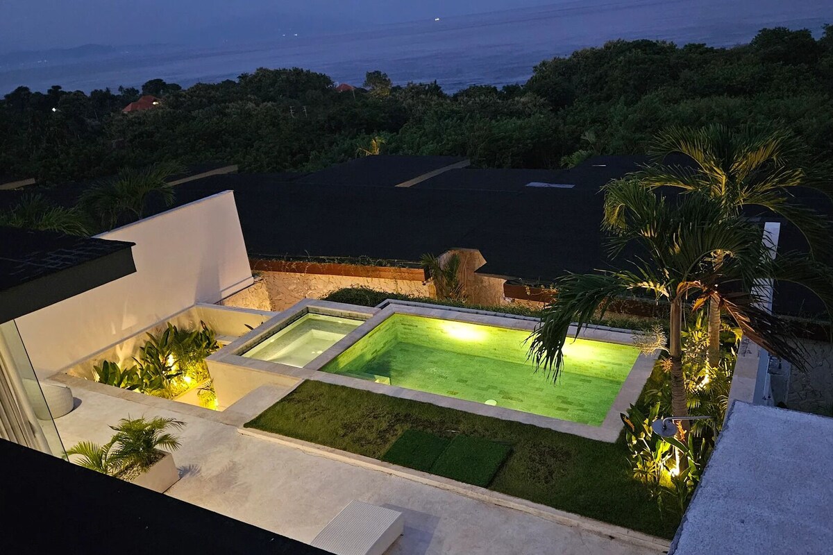 A rooftop pool with a view of the ocean, surrounded by greenery and modern architecture.