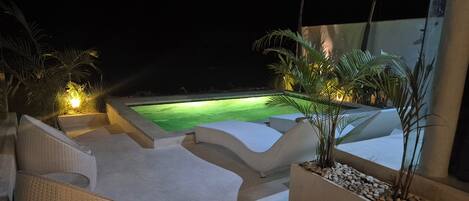A nighttime outdoor area with a swimming pool, lounge chairs, and potted plants.