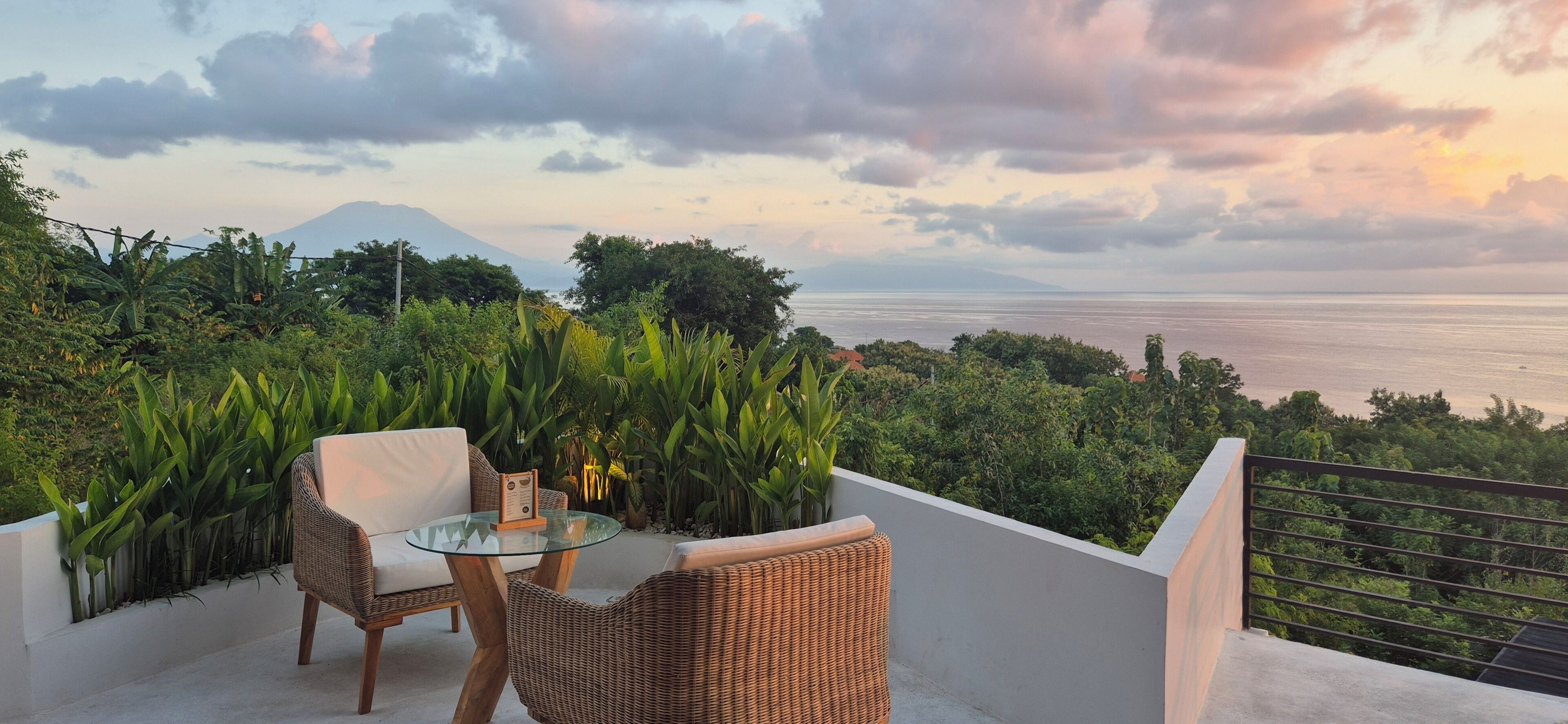 A balcony with wicker furniture, a glass table, and a view of the sea and a mountain.