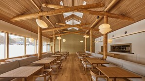 Un vaste espace de restauration intérieur, doté de tables et de bancs en bois, de grandes fenêtres et d’un haut plafond avec des poutres apparentes.