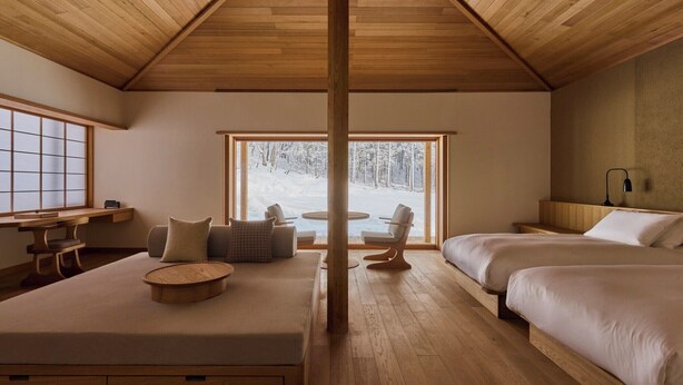 Une pièce chaleureuse avec un plafond en bois, une grande fenêtre donnant sur une forêt enneigée, un canapé, une table ronde et deux lits.