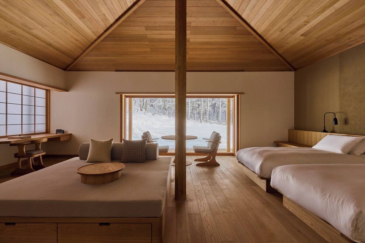 Ein gemütliches Zimmer mit Holzbalkendecke, einem großen Fenster mit Blick auf einen verschneiten Wald, einer Couch, einem runden Tisch und zwei Betten.