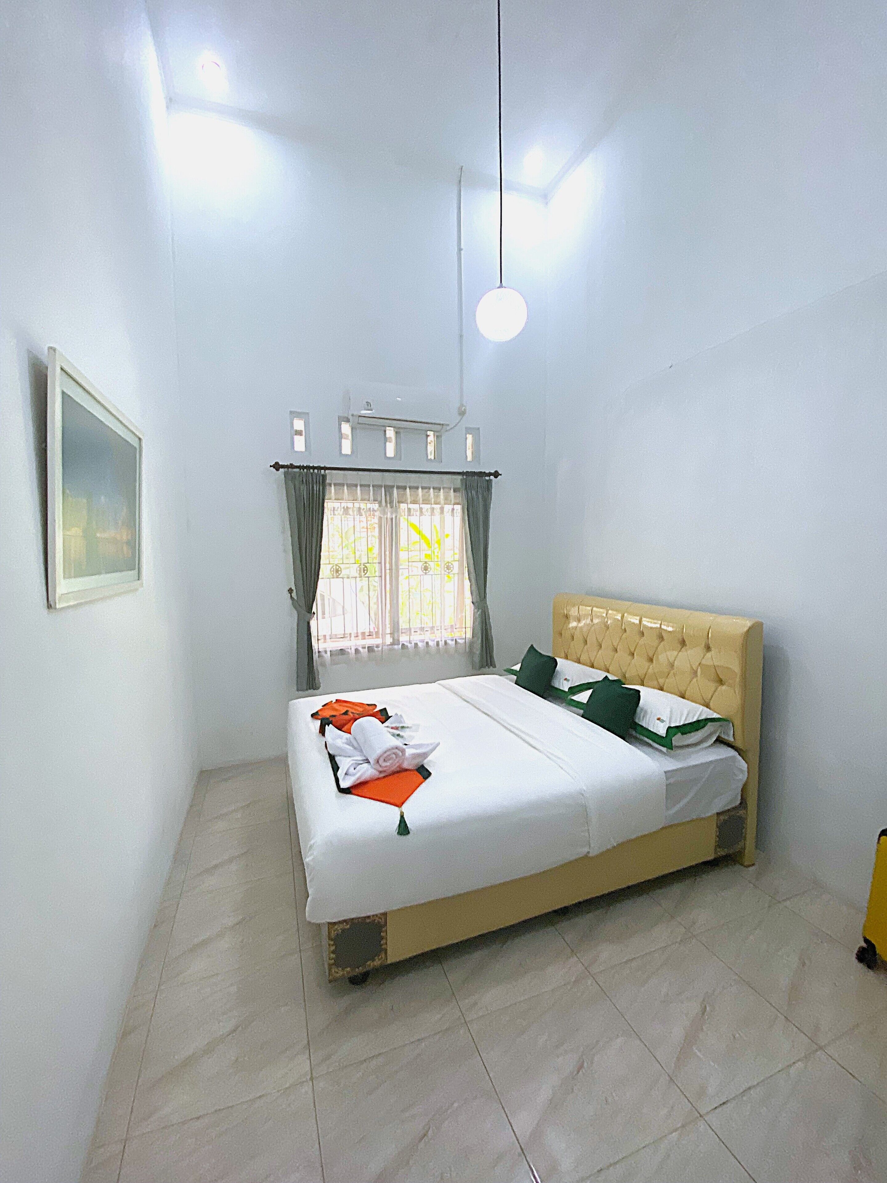 Cama bem arrumada com lençóis brancos, um travesseiro verde e detalhes em laranja. Há uma janela com cortinas, um lustre pendurado e um quadro na parede.