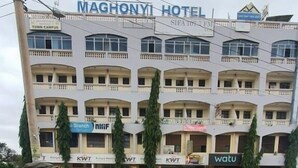 A multi-story building with the sign "MAGHONYI HOTEL" at the top. There are several storefronts with signs like "KWFT" and "watu" on the ground floor. A pickup truck is parked in front.